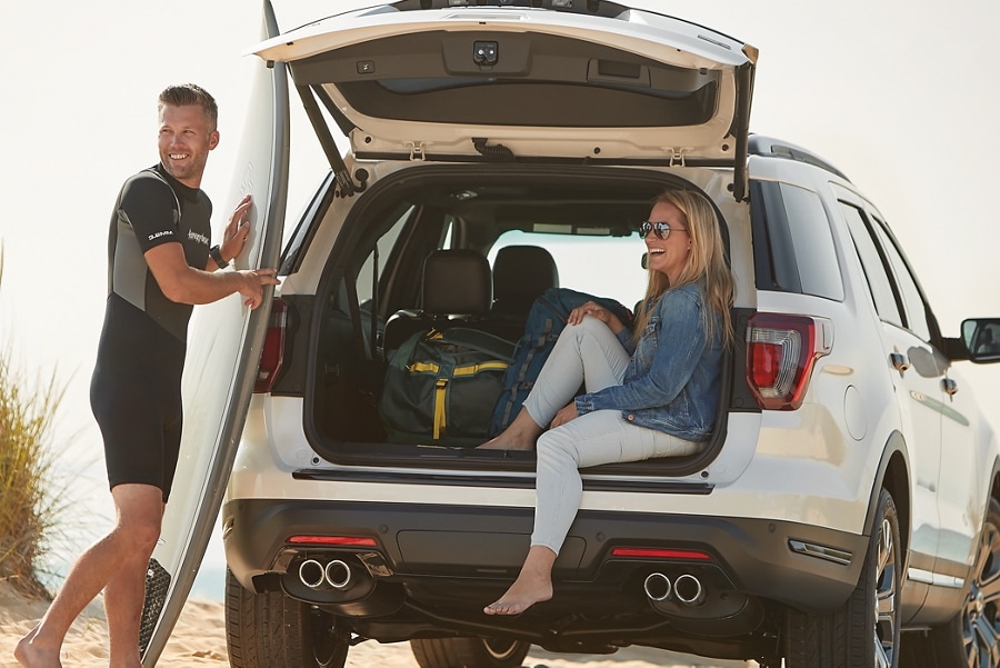 Couple with a surfboard sitting in the back of a parked Ford SUV.